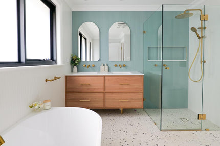 Modern bathroom featuring a floating timber vanity with double basins, arched mirrors, a frameless glass shower with brushed brass fixtures, a freestanding white bathtub, terrazzo flooring, and soft mint-green wall tiles in a bright, contemporary space.