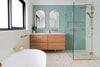 Modern bathroom featuring a floating timber vanity with double basins, arched mirrors, a frameless glass shower with brushed brass fixtures, a freestanding white bathtub, terrazzo flooring, and soft mint-green wall tiles in a bright, contemporary space.
